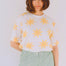 Woman wearing a white shirt with yellow sun patterns and light green pants against a white wall.