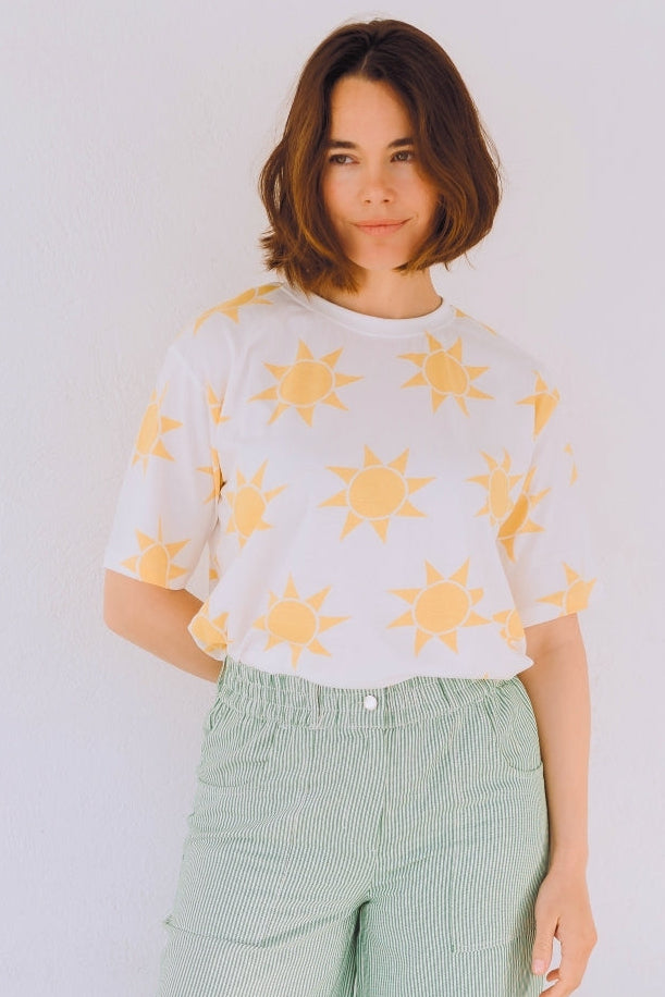 Woman wearing a white shirt with yellow sun patterns and light green pants against a white wall.