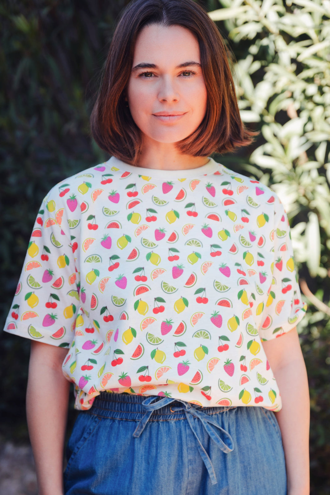 A person wearing a white relaxed fit t-shirt with a fruit salad print, including various colorful fruits such as strawberries and grapes, paired with blue denim shorts.