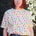 A person wearing a white relaxed fit t-shirt with a fruit salad print, including various colorful fruits such as strawberries and grapes, paired with blue denim shorts.