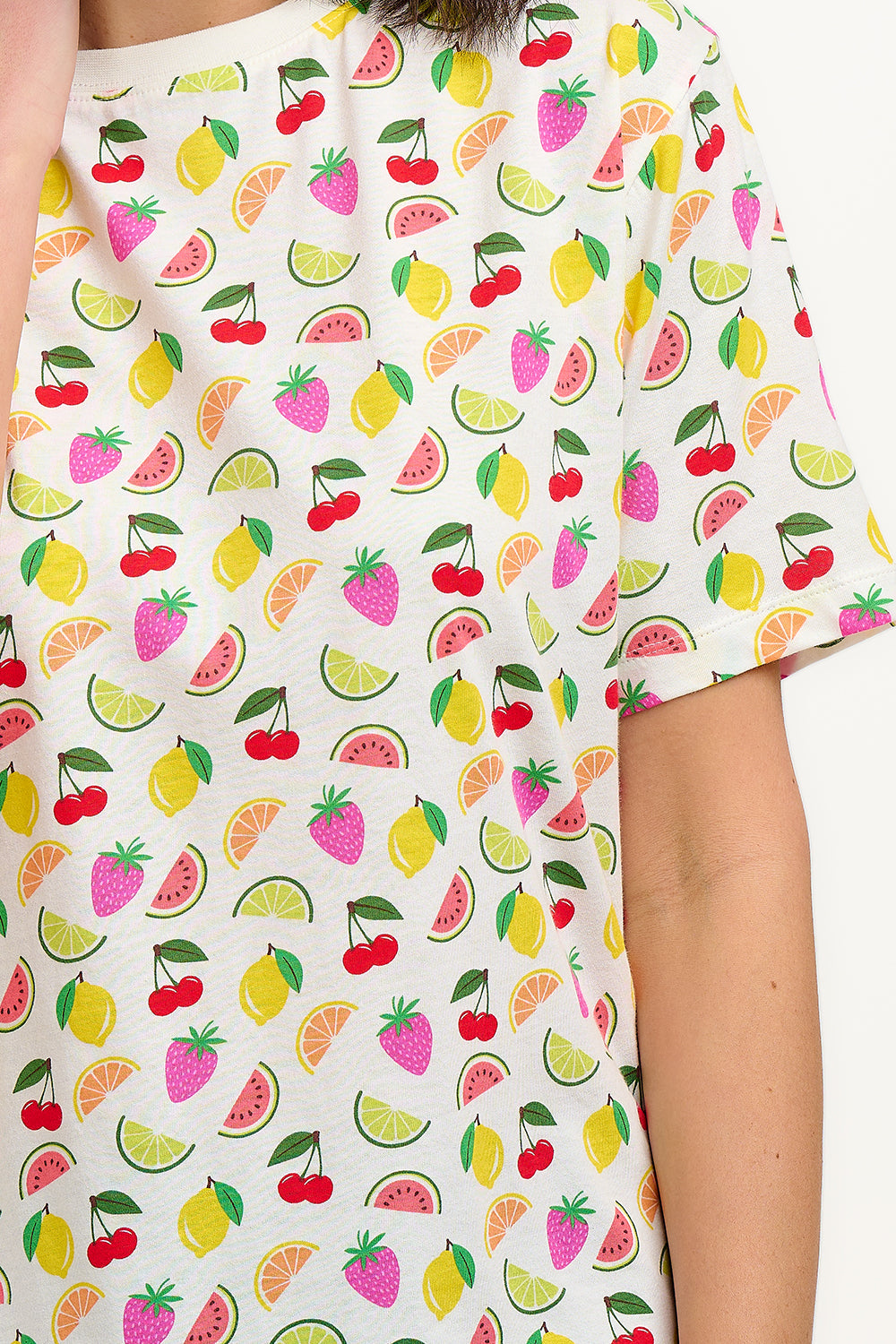 A person wearing a white relaxed fit t-shirt with a fruit salad print, including various colorful fruits such as strawberries and grapes. 