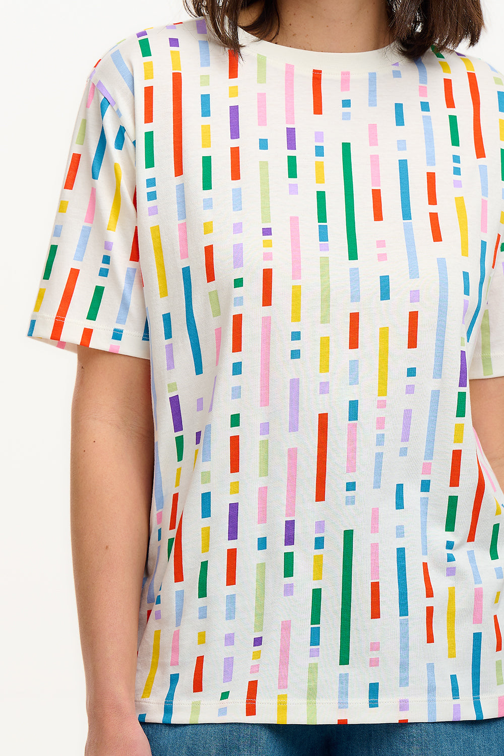 A woman smiling and wearing an oversized white t-shirt with a broken lines multicolor print, paired with denim shorts.