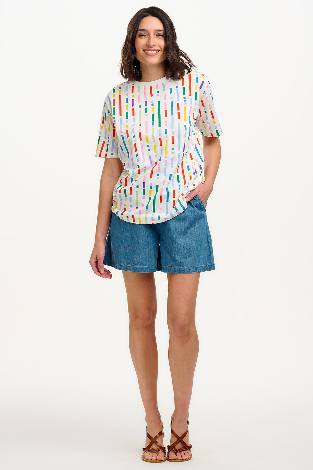 A woman smiling and wearing an oversized white t-shirt with a broken lines multicolor print, paired with denim shorts.