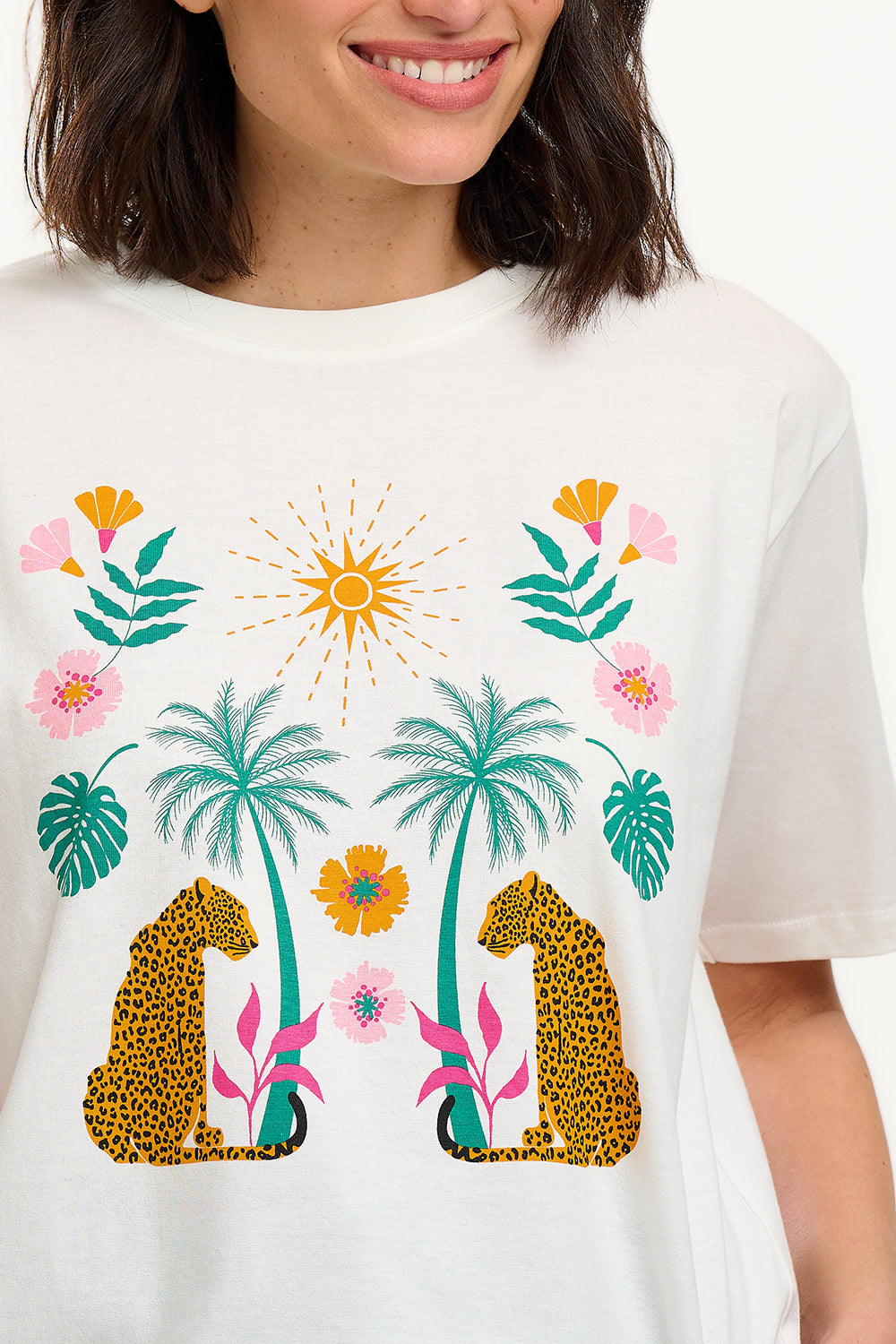 A woman smiling and wearing an off-white colored t-shirt with a mirrored jungle print design, featuring palm trees, leopards, and flowers.