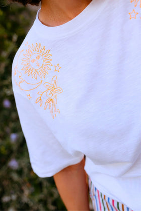 White t-shirt with yellow embroidered designs on a blurred natural background