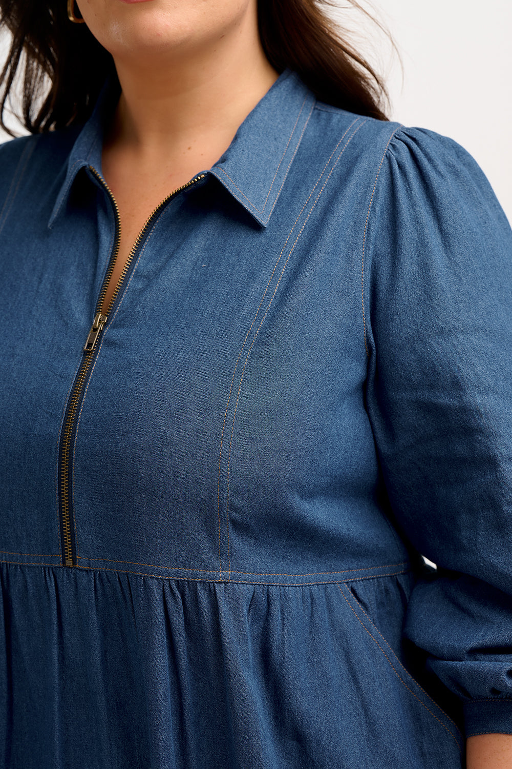 A mid-length denim smock dress with a front zip fastening and cuffed sleeves, worn by a model posing against a white background.