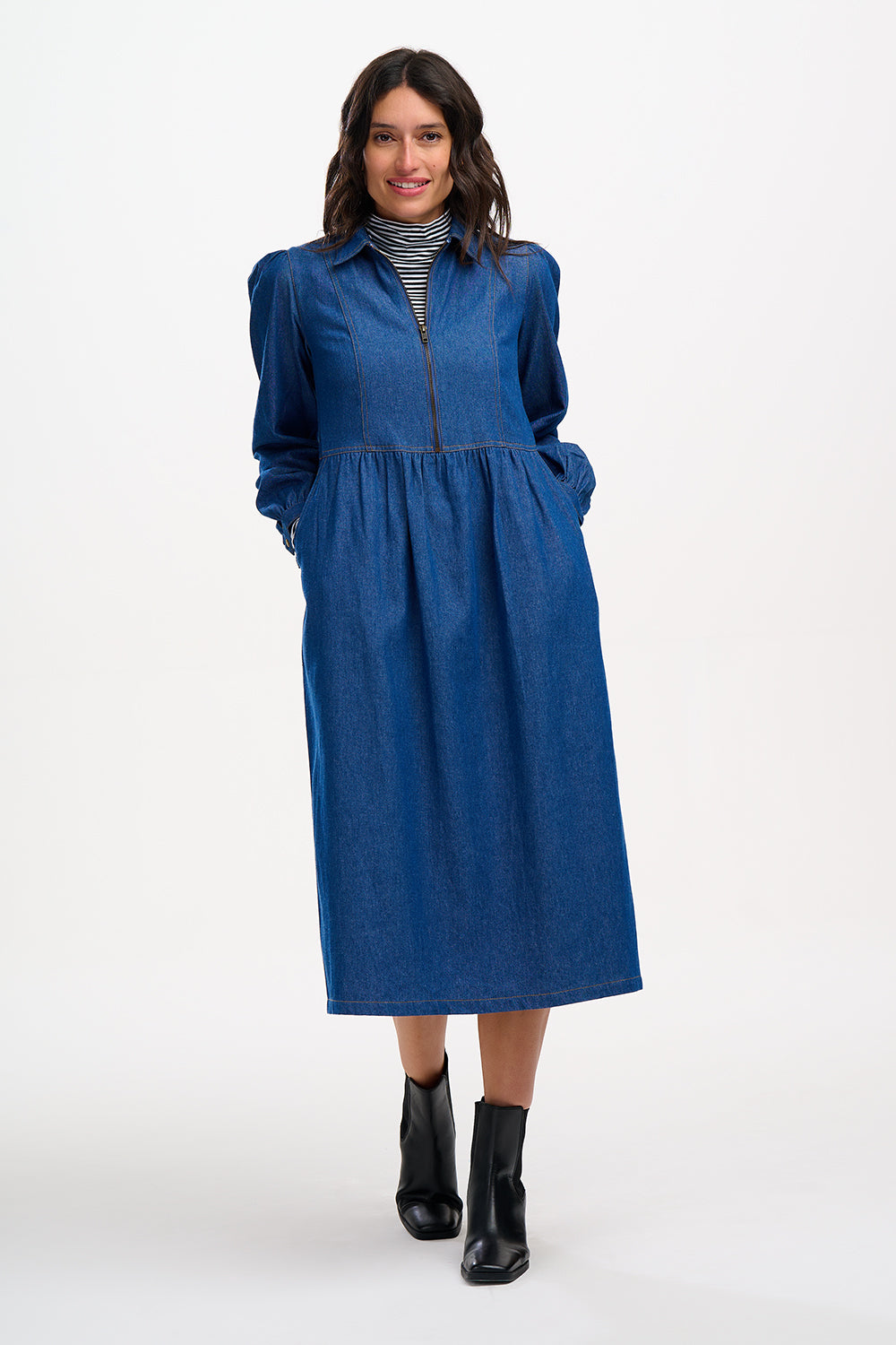 A woman standing, wearing a mid-blue denim smock dress with an exposed front zip, blouson cuffed sleeves, and a midi length skirt, paired with black ankle boots.