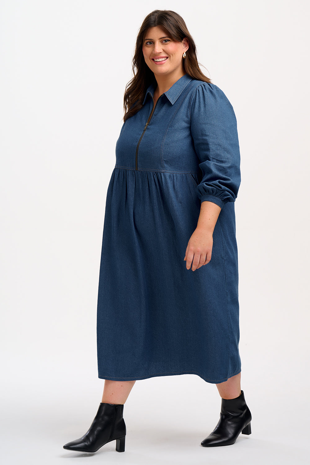 A mid-length denim smock dress with a front zip fastening and cuffed sleeves, worn by a model posing against a white background.