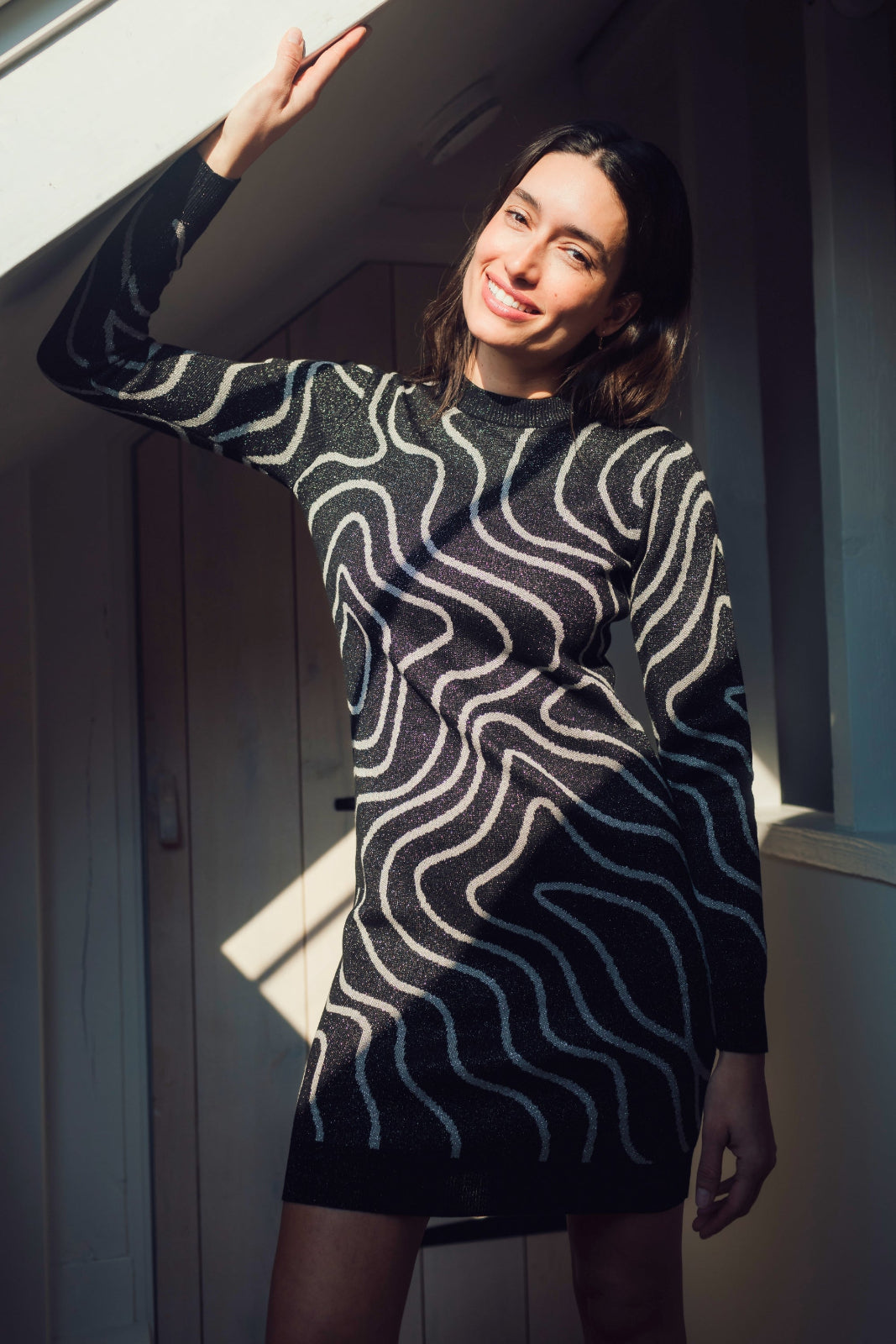 Woman wearing a black and white patterned dress standing in a room with sunlight filtering through.