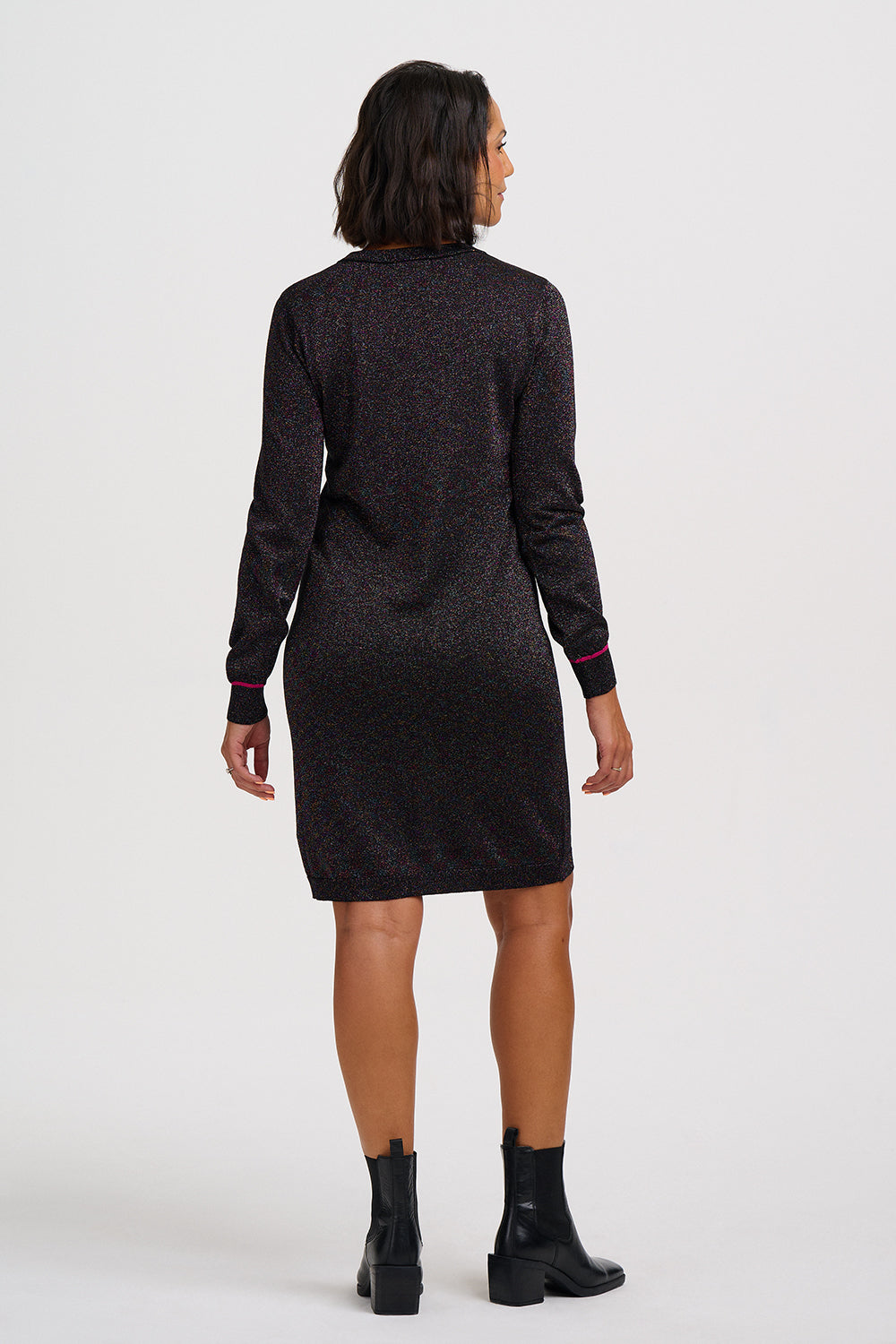 A woman viewed from the back wearing a dark long-sleeve knit dress with a subtle speckled texture and black ankle boots, against a plain light gray studio background.