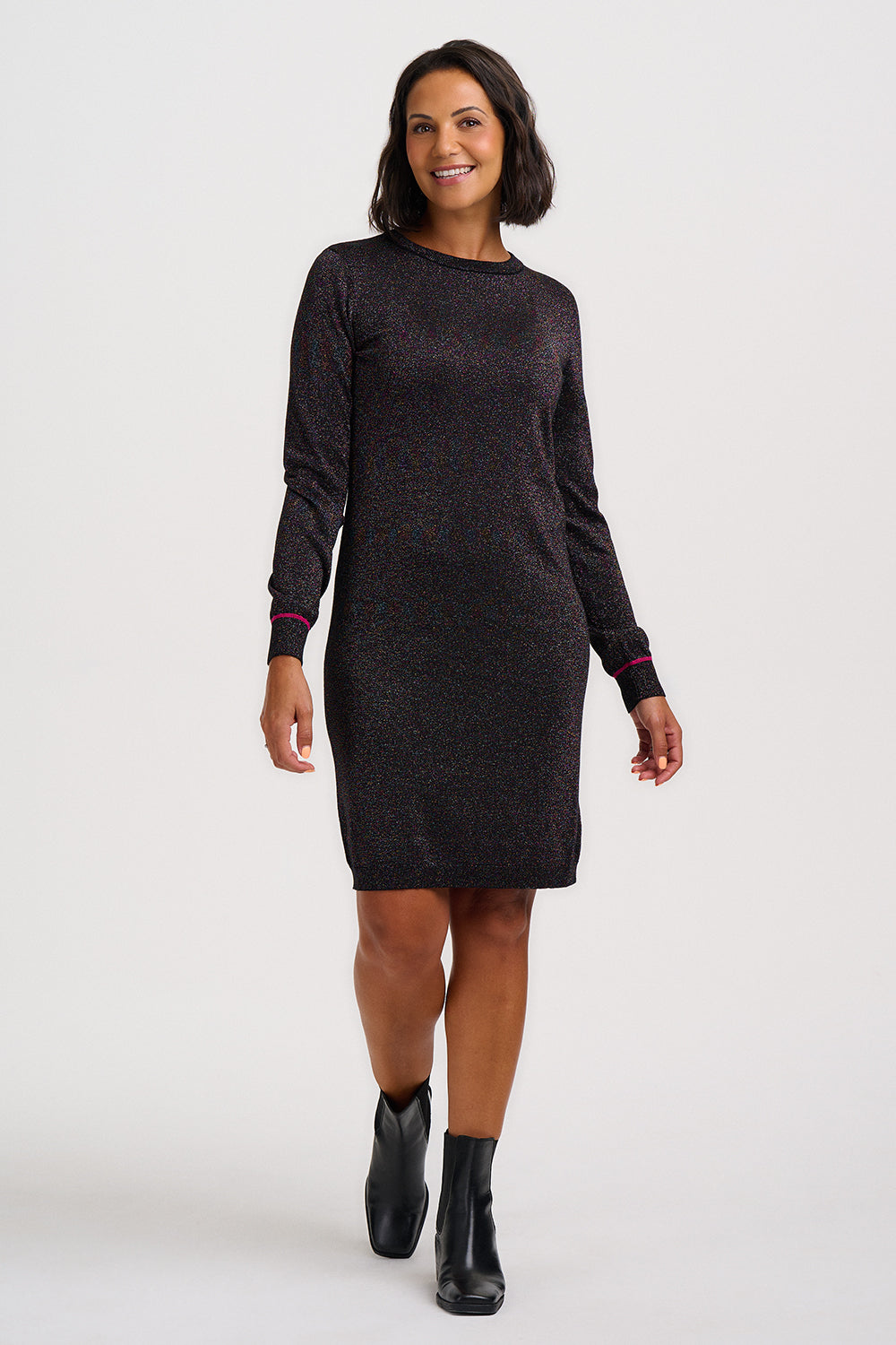 A woman modeling a long-sleeve knit dress with a dark charcoal and black micro-spotted pattern, featuring pink accent lines on the cuffs, paired with black ankle boots against a light gray studio background.