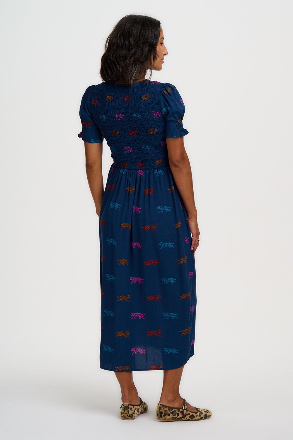 A woman is shown from the back wearing a navy blue midi dress with short puff sleeves and an allover colorful dragonfly print. She is standing against a plain white studio background.