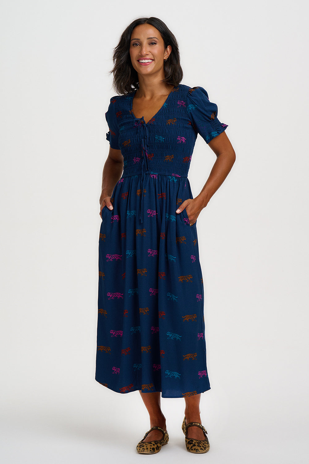 A woman modeling a navy blue midi dress with a colorful butterfly pattern, featuring puff sleeves, a V-neckline, and side pockets, set against a plain light gray background.