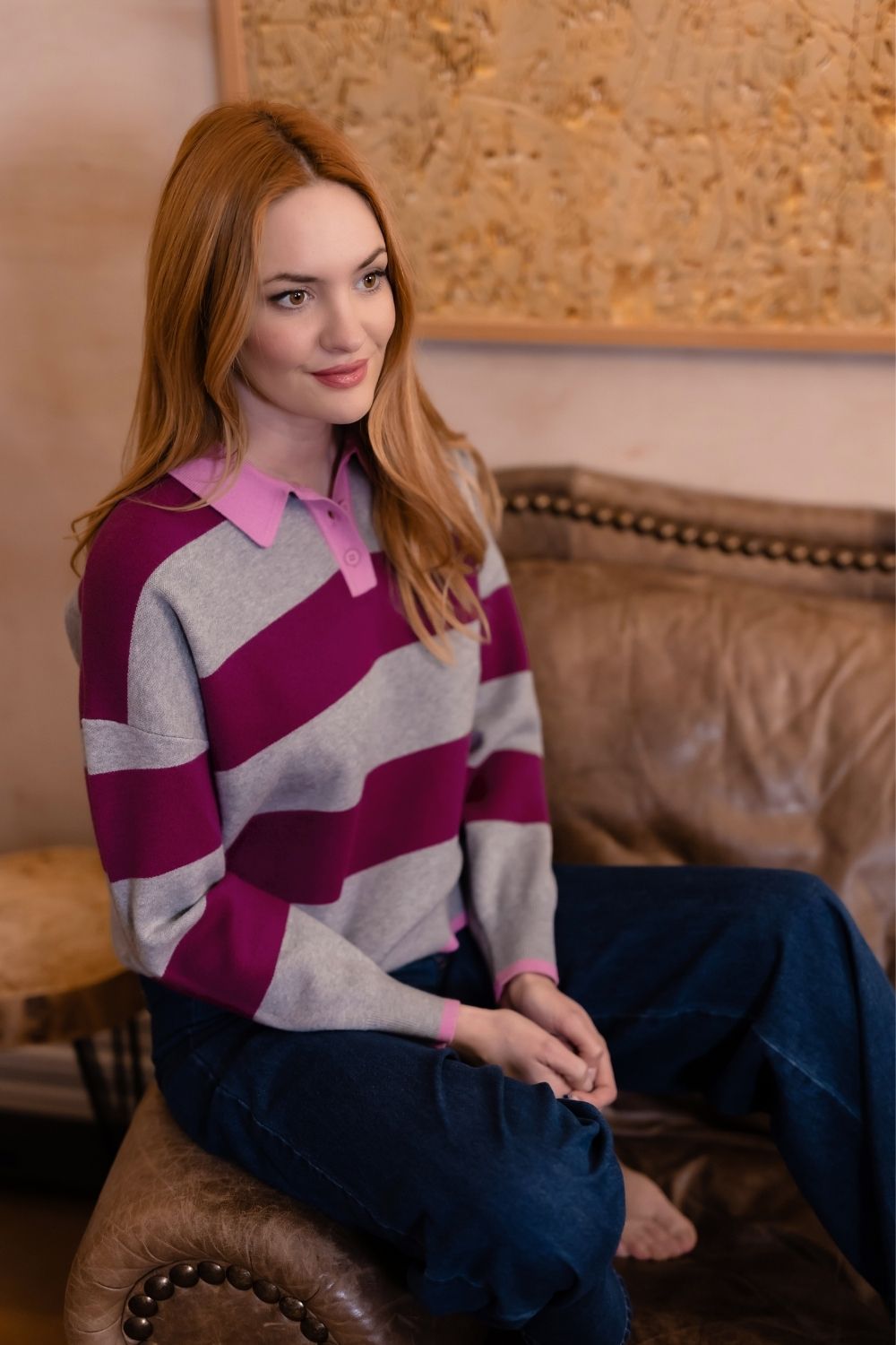 A woman with red hair sits on a brown leather couch wearing a gray and magenta striped long-sleeve polo sweater with a pink collar, paired with dark blue jeans.