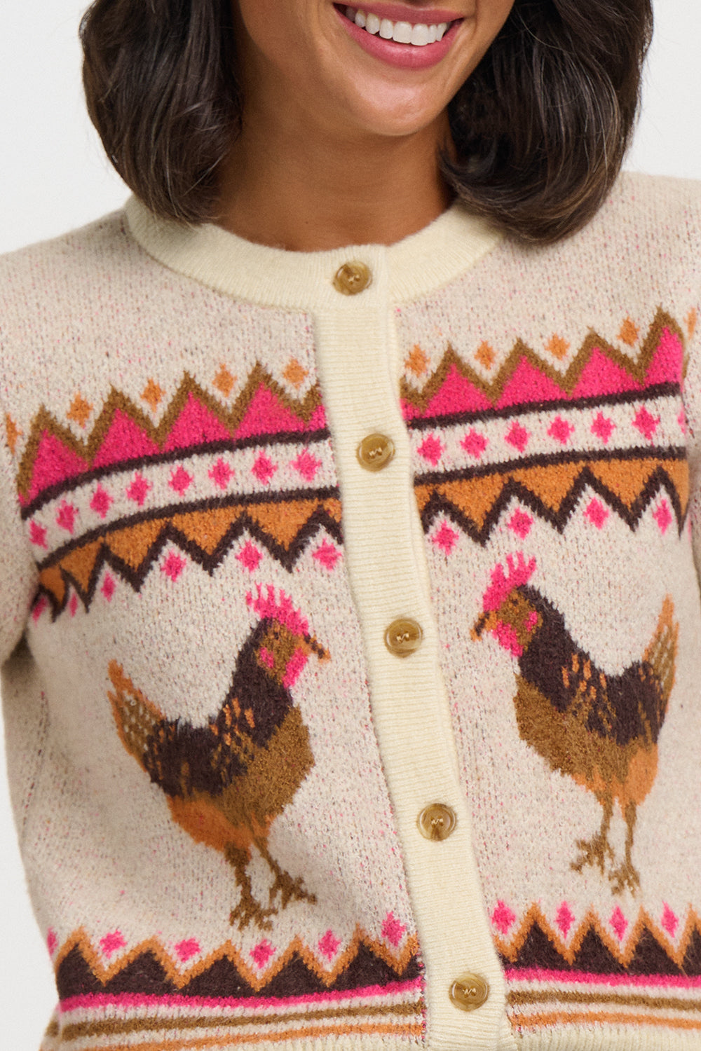 Close-up of a cream knit cardigan sweater with a colorful geometric pattern and two brown chickens, featuring gold-tone buttons, worn by a model.