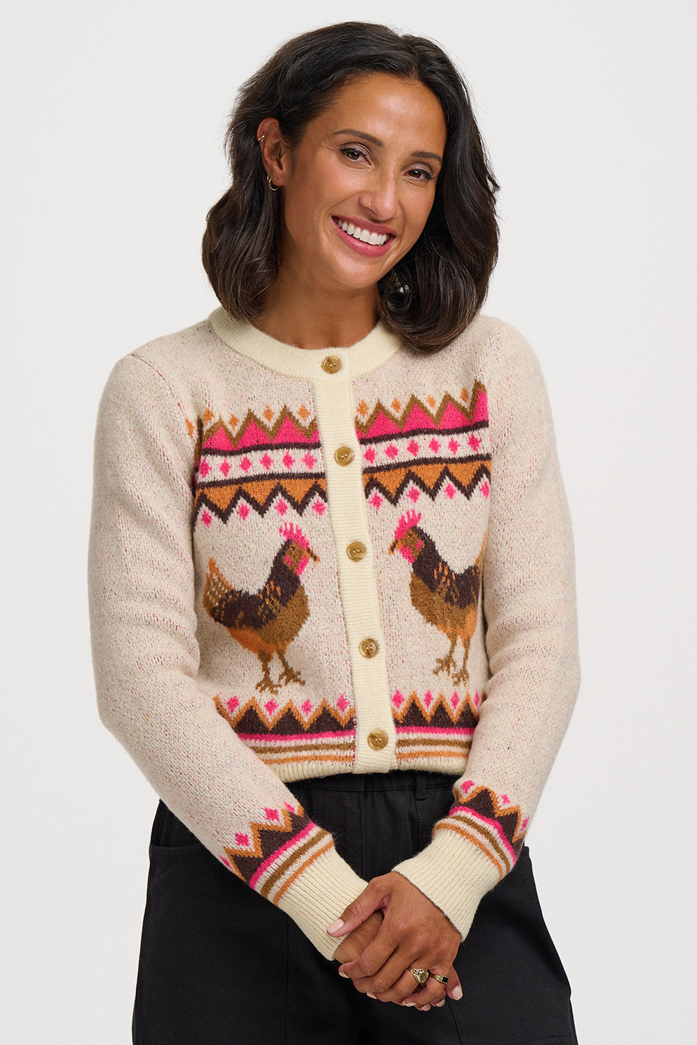 A woman wearing a cream-colored knit cardigan sweater with a colorful geometric pattern and two brown rooster illustrations on the front, paired with black pants, against a light gray studio background.