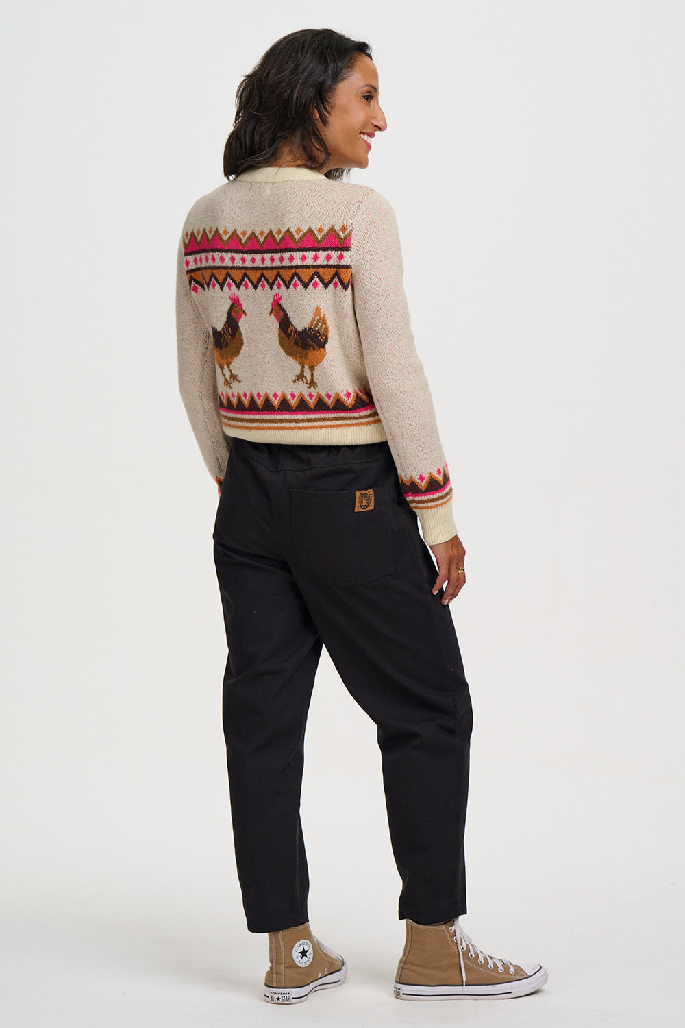 A woman shown from the back wearing a cream knit sweater with a colorful geometric pattern and two rooster illustrations across the upper back, paired with black pants and tan high-top sneakers, against a plain light gray studio background.