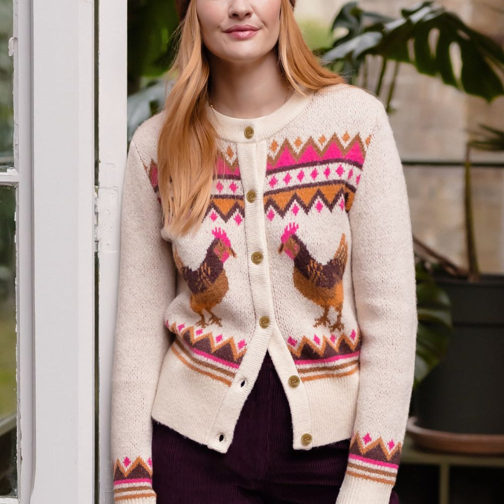 A woman wearing a cream-colored knit cardigan with a colorful geometric pattern and two brown chickens, paired with a brown beanie and dark purple pants, standing in front of a white window frame.