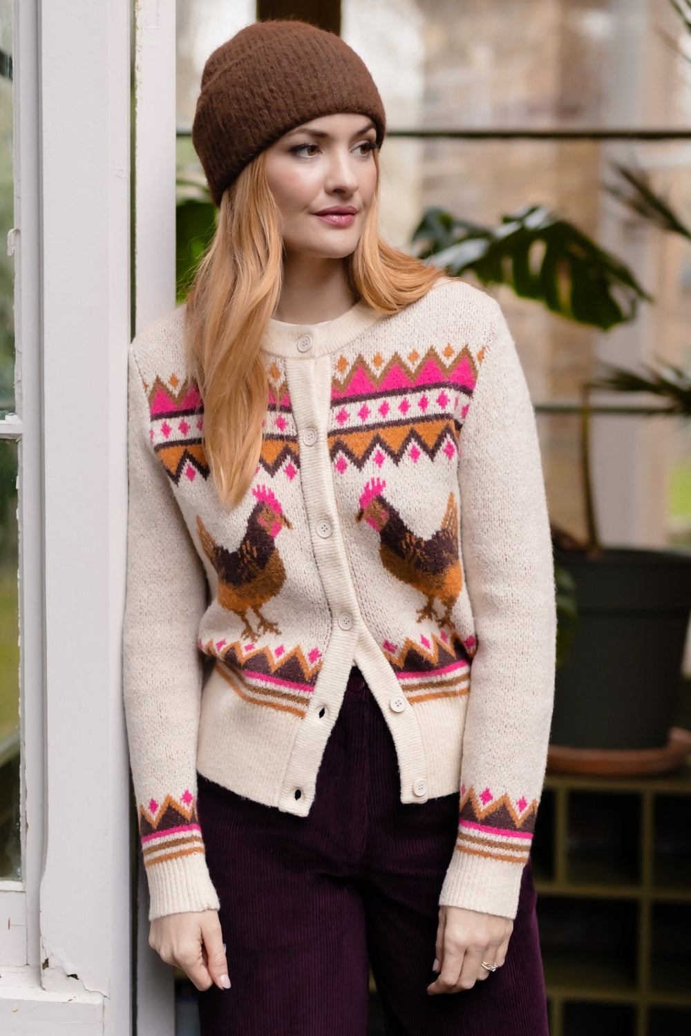 A woman wearing a cream-colored knit cardigan with a colorful Fair Isle pattern featuring chickens and geometric borders, a brown beanie, and dark purple trousers, standing next to a white window frame.