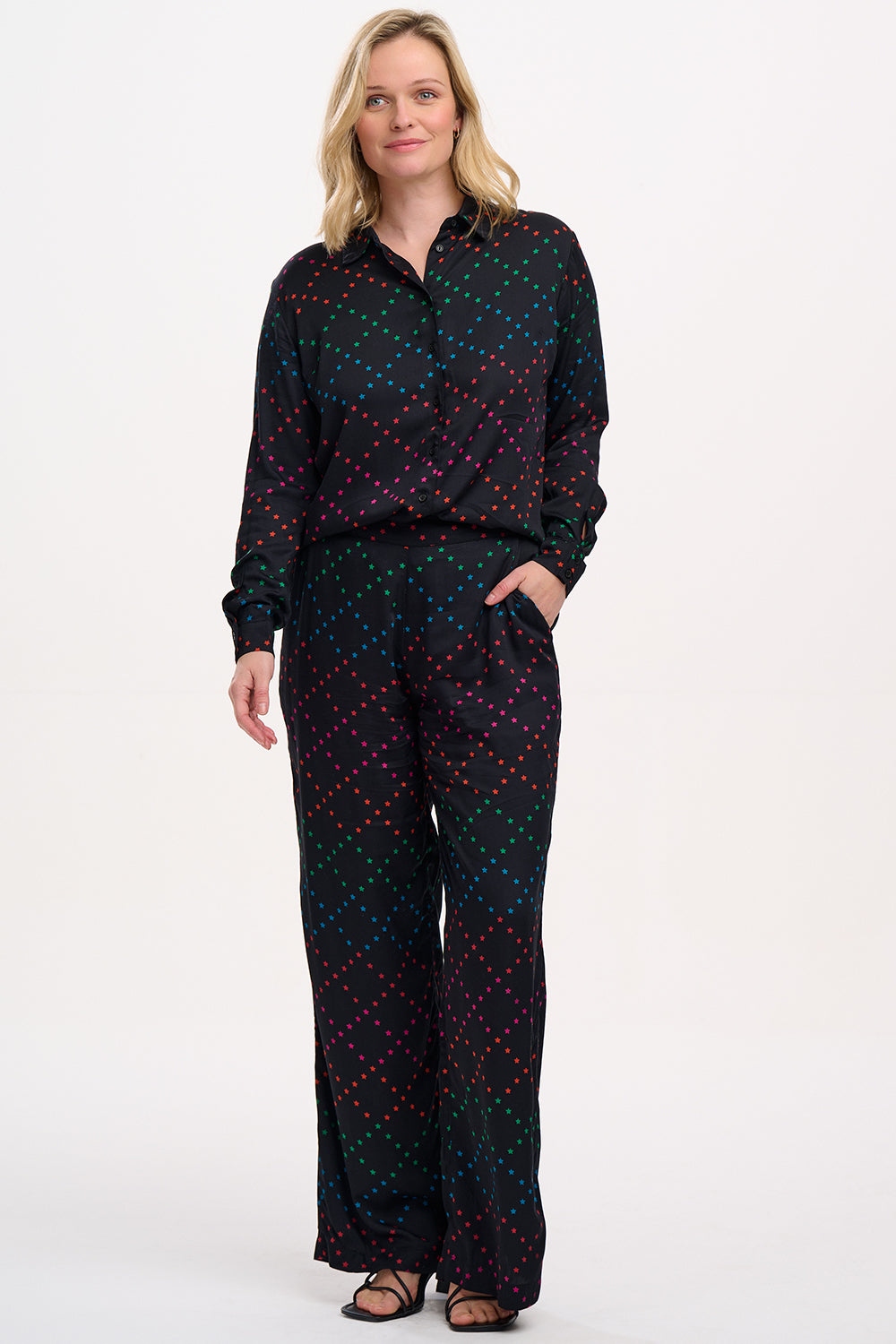 A person standing wearing black trousers with a colorful geometric star pattern on the lower half, paired with a black top and sandals.