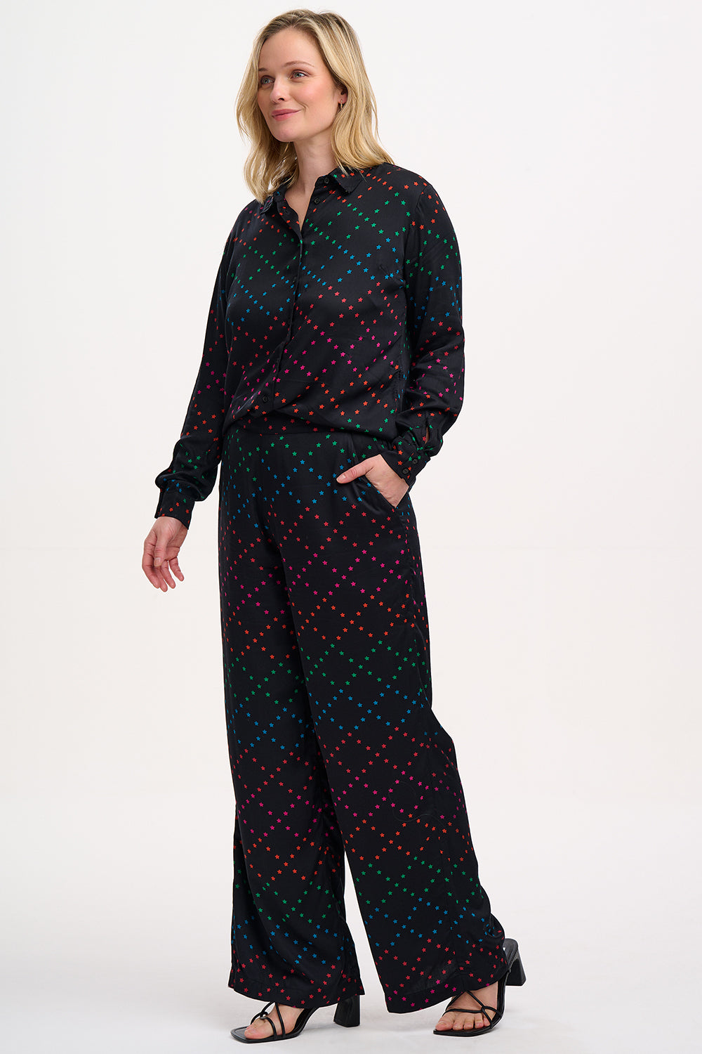 A person standing wearing black trousers with a colorful geometric star pattern on the lower half, paired with a black top and sandals.