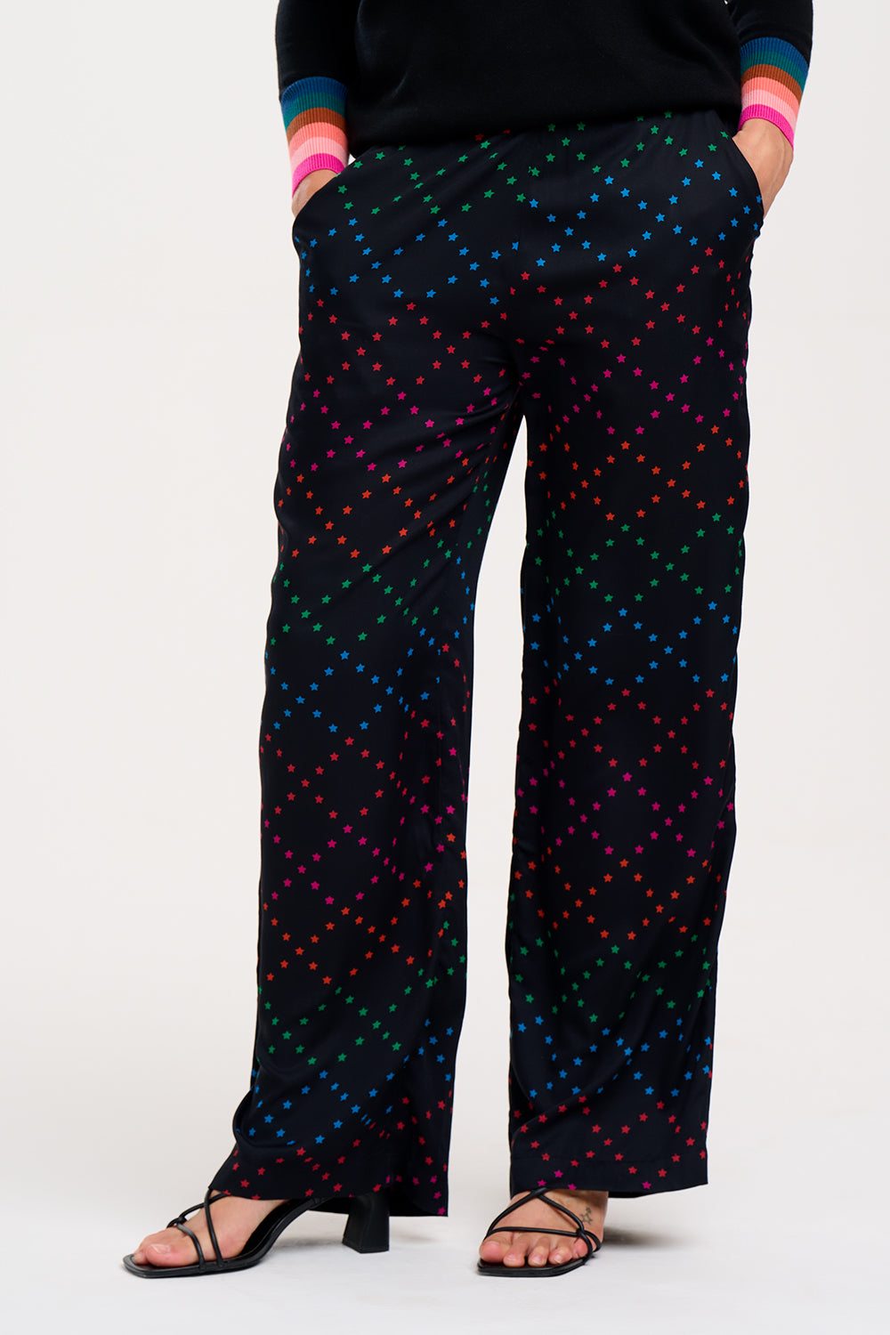 A person standing wearing black trousers with a colorful geometric star pattern on the lower half, paired with a black top and sandals.