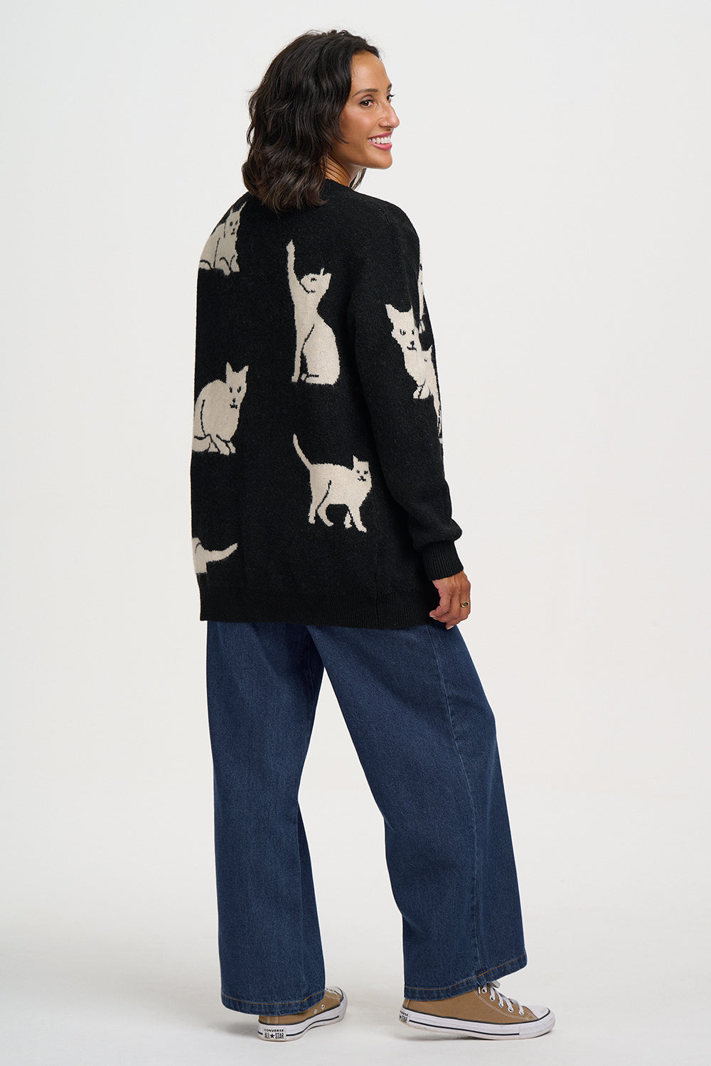 A woman wearing a black knit sweater with a white cat pattern, paired with wide-leg blue jeans and brown sneakers, shown from a side-back angle against a plain white studio background.