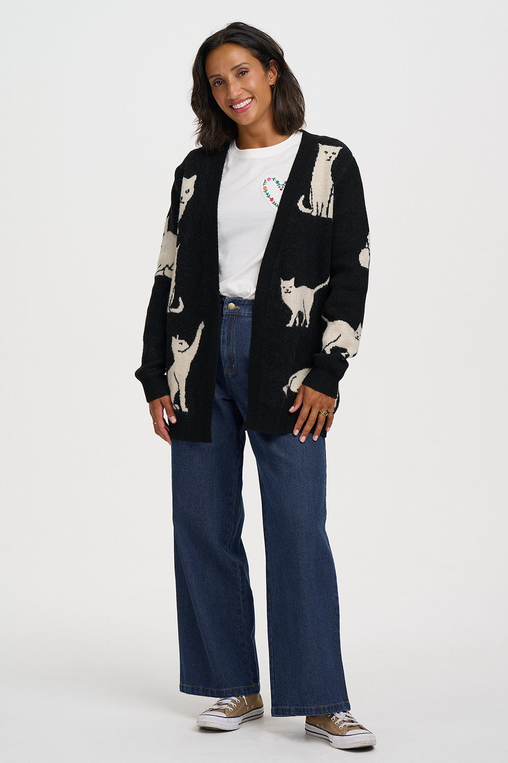A woman modeling a black knit cardigan with cream cat illustrations, worn over a white t-shirt and blue jeans, against a light gray studio background.