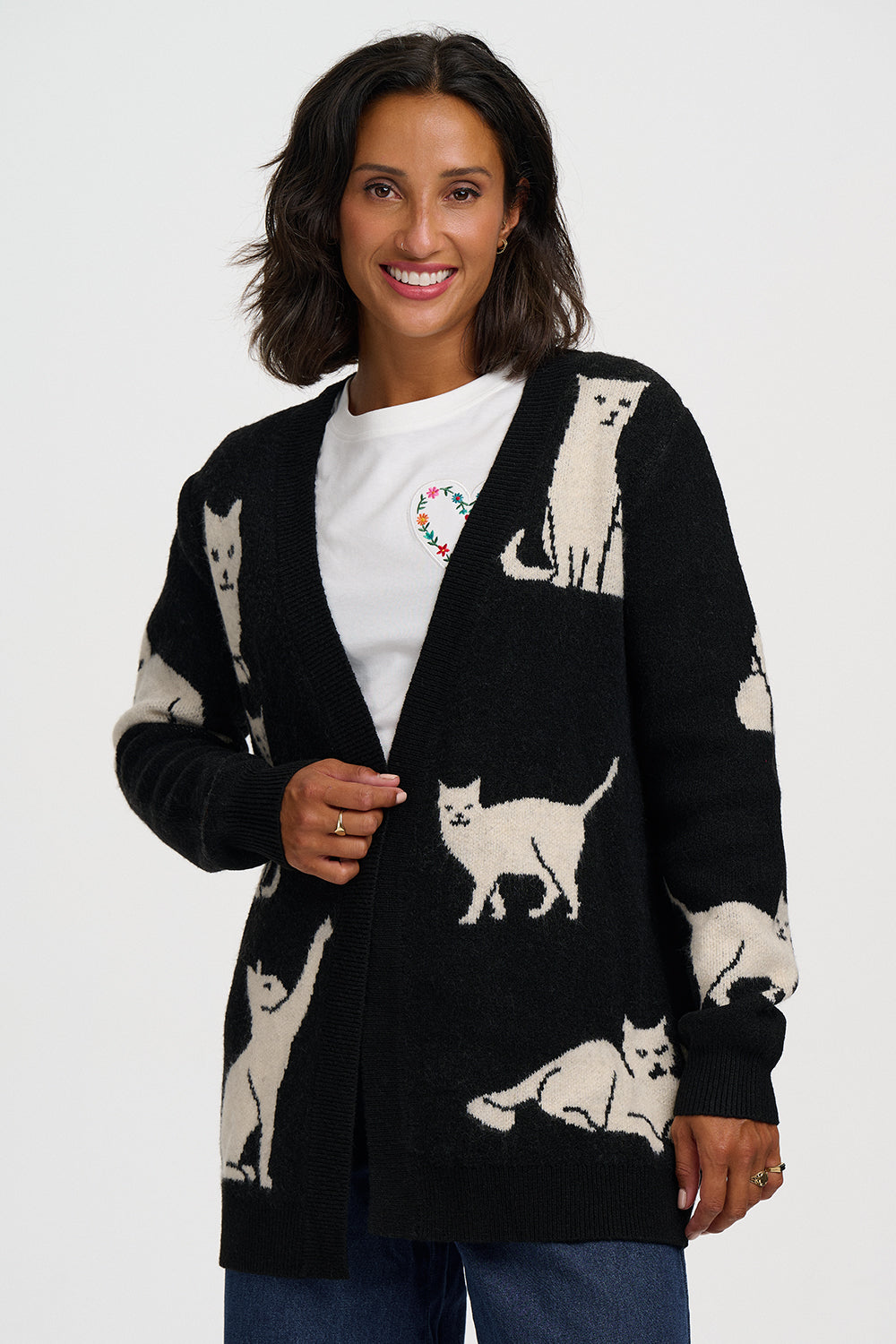 A woman wearing a black open-front cardigan with cream-colored cat illustrations, layered over a white graphic t-shirt and blue jeans, against a plain light gray background.