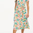 A woman wearing an off-white shirt dress with a colorful floral print, featuring a button-up front, a shirt collar, and a tie waist belt.