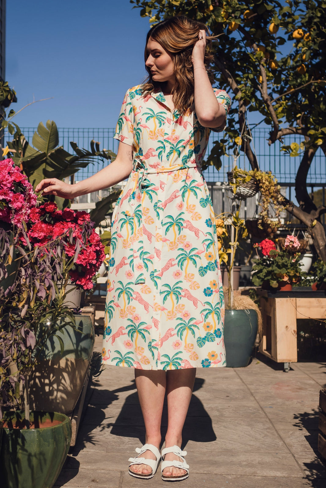 A woman standing in a garden wearing an off-white shirt dress with a colorful floral print, featuring a button-up front, a shirt collar, and a tie waist belt.
