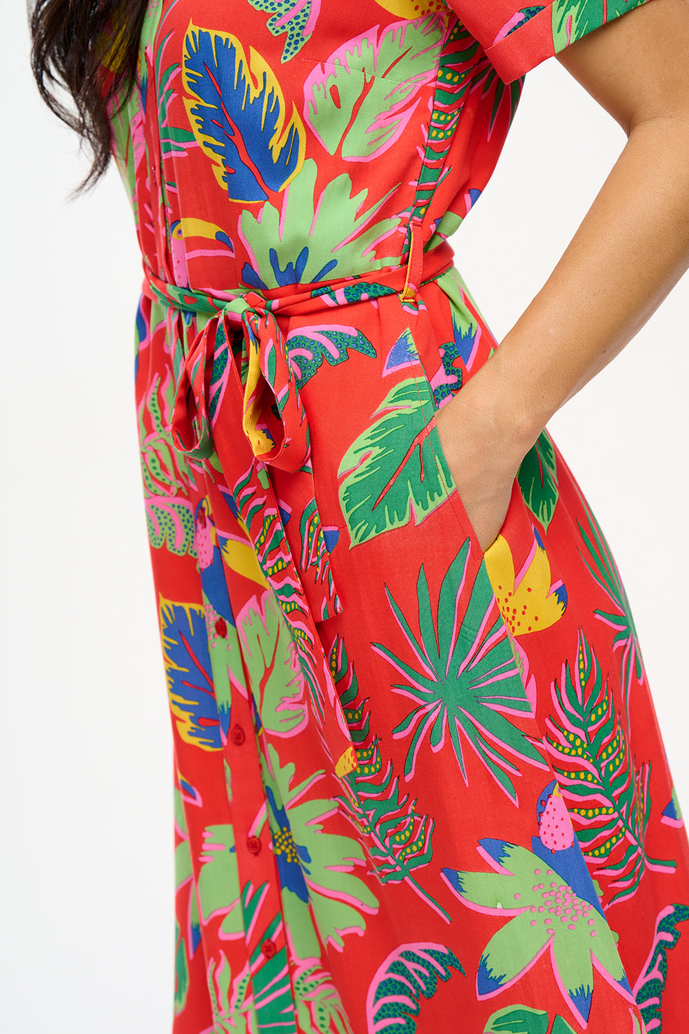 A woman wearing a red shirt dress with a Tropical Toucans print, featuring a button-up front, a shirt collar, and a tie waist belt.