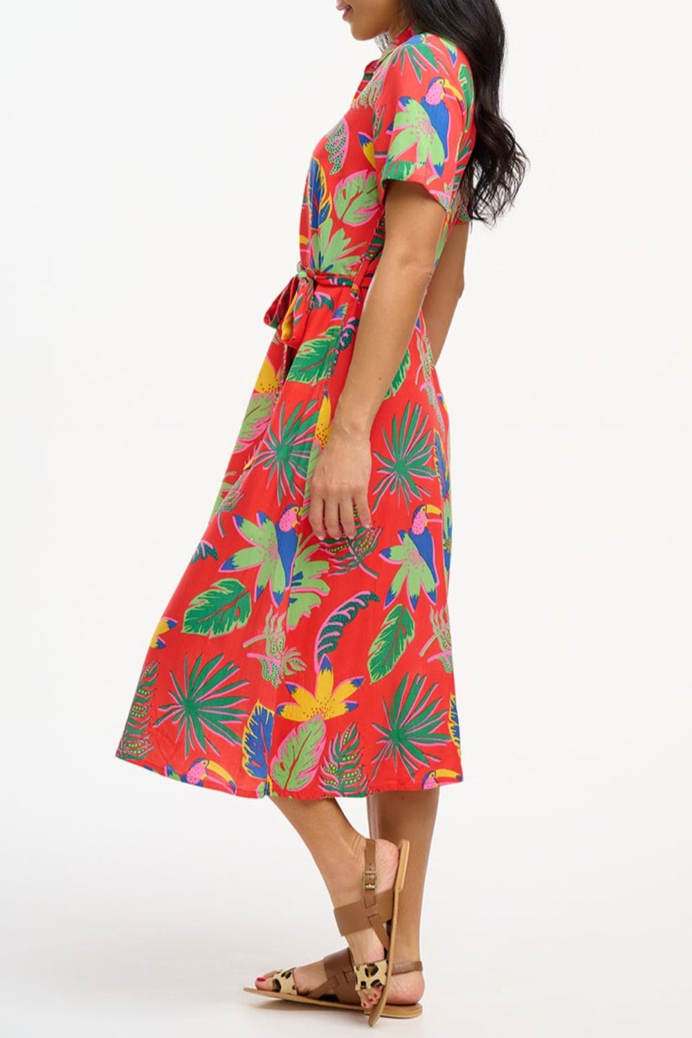 A woman wearing a red shirt dress with a Tropical Toucans print, featuring a button-up front, a shirt collar, and a tie waist belt.