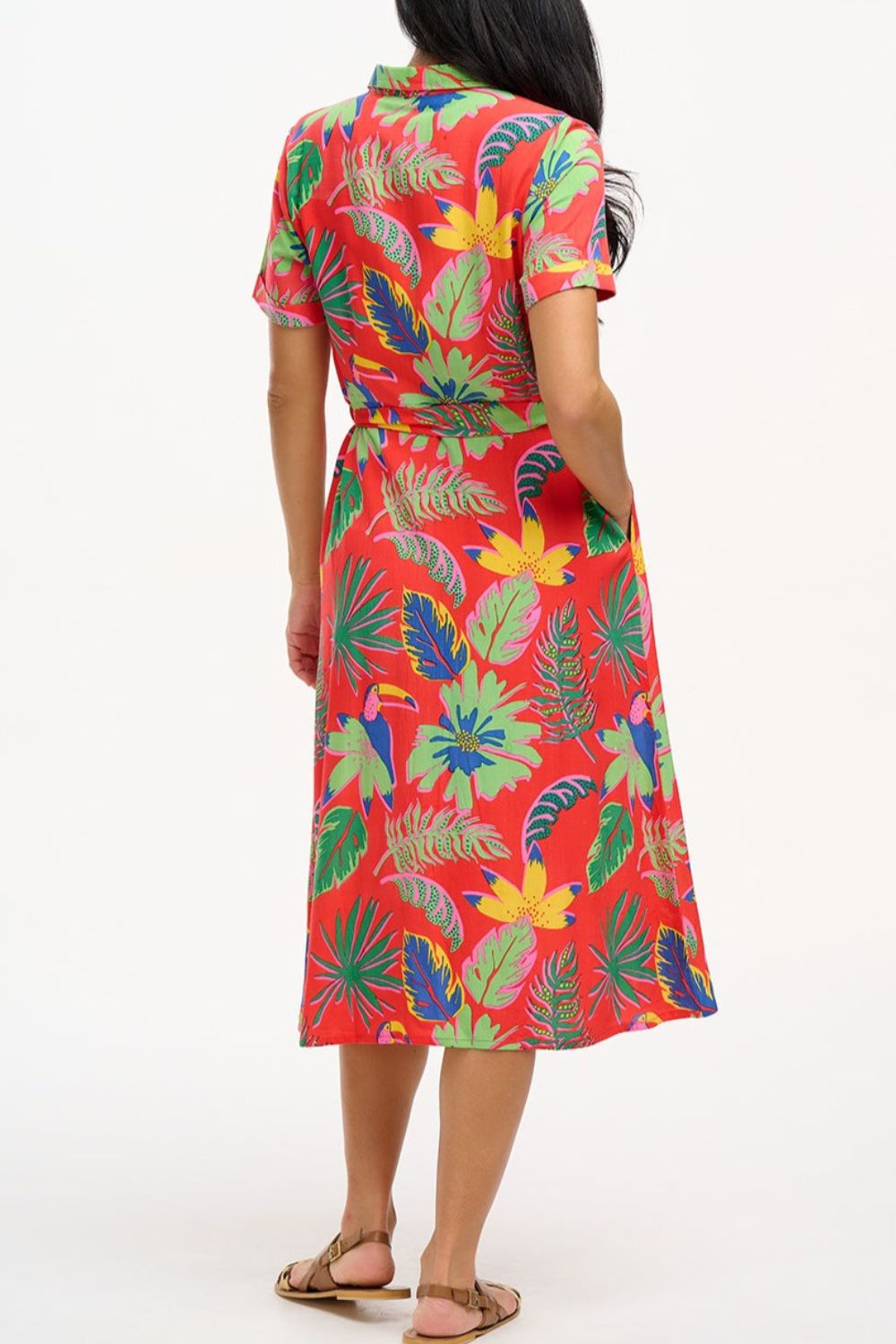 A woman wearing a red shirt dress with a Tropical Toucans print, featuring a button-up front, a shirt collar, and a tie waist belt.
