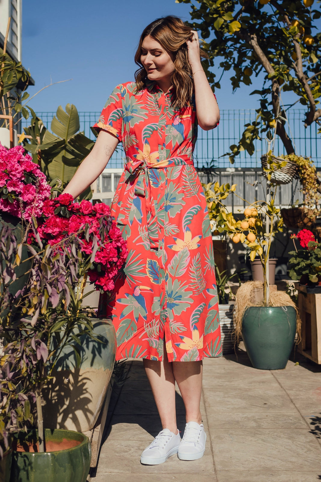 A woman standing in a garden wearing a red shirt dress with a Tropical Toucans print, featuring a button-up front, a shirt collar, and a tie waist belt.