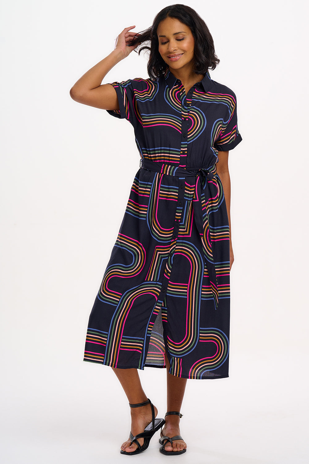 A model wearing a midi length shirt dress with a traditional shirt collar, button placket, and a full skirt with an elasticated waist. The dress has a black base with a 70s-inspired geometric retro print in red and pink.
