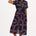 A model wearing a midi length shirt dress with a traditional shirt collar, button placket, and a full skirt with an elasticated waist. The dress has a black base with a 70s-inspired geometric retro print in red and pink.