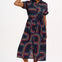 A model wearing a midi length shirt dress with a traditional shirt collar, button placket, and a full skirt with an elasticated waist. The dress has a black base with a 70s-inspired geometric retro print in red and pink.