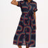 A model wearing a midi length shirt dress with a traditional shirt collar, button placket, and a full skirt with an elasticated waist. The dress has a black base with a 70s-inspired geometric retro print in red and pink.