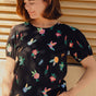 A model wearing a black jersey t-shirt with a floral and hummingbird print, round neckline, and short puffed sleeves.