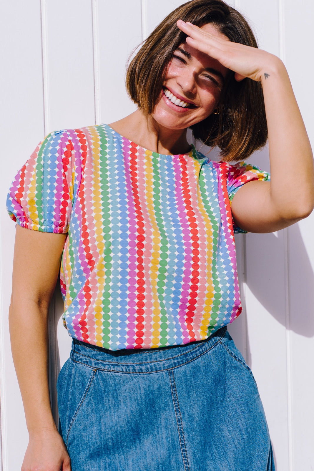 A woman smiling and posing in a multicolored jersey top with a geometric pattern of rainbow circles, paired with a denim skirt.