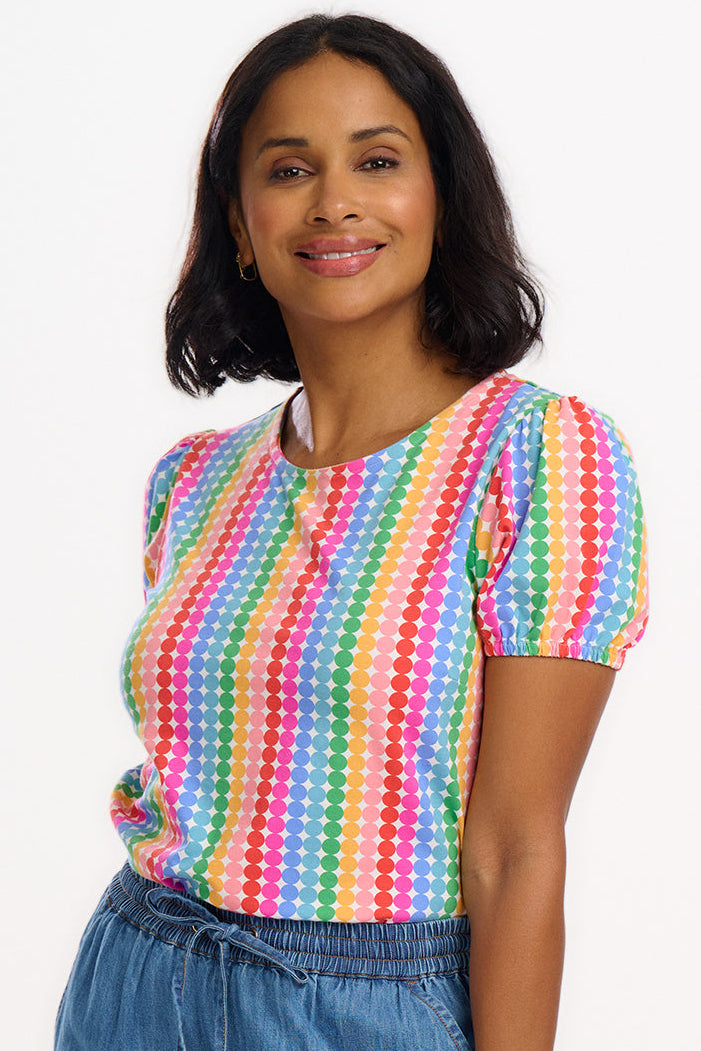 A woman wearing a multicolored jersey top with a pattern of rainbow circles on an off-white base and short puff sleeves, paired with blue jeans.