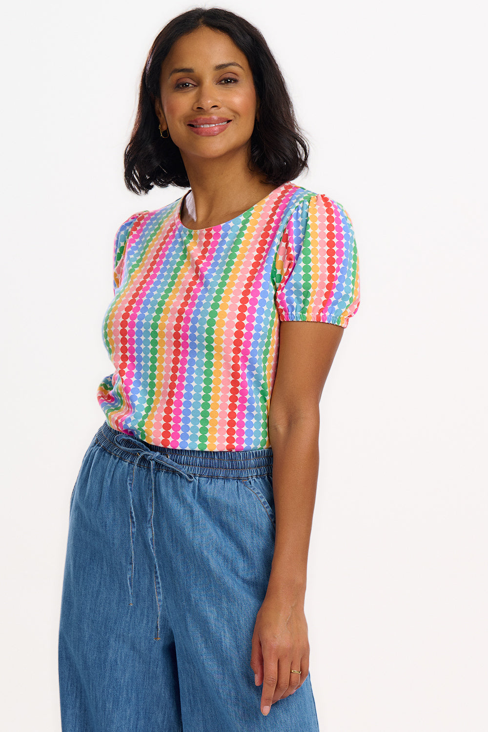 A woman wearing a multicolored jersey top with a pattern of rainbow circles on an off-white base and short puff sleeves, paired with blue jeans.