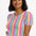 A woman wearing a multicolored jersey top with a pattern of rainbow circles on an off-white base and short puff sleeves, paired with blue jeans.