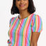 A woman wearing a multicolored jersey top with a pattern of rainbow circles on an off-white base and short puff sleeves, paired with blue jeans.