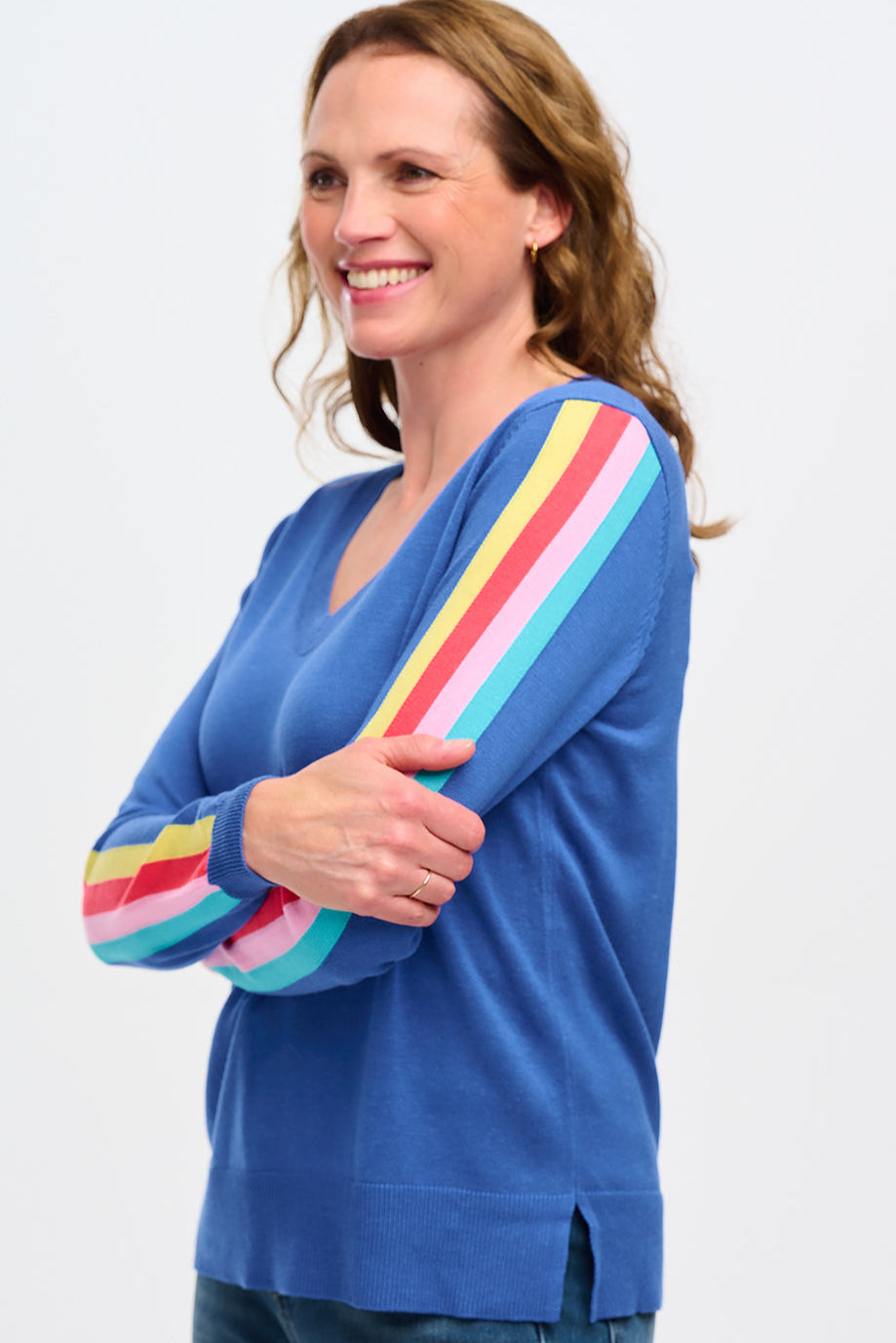 A woman wearing a blue V-neck jumper with colorful stripes on the sleeves. The jumper has a semi-fitted silhouette and falls to mid-hip.