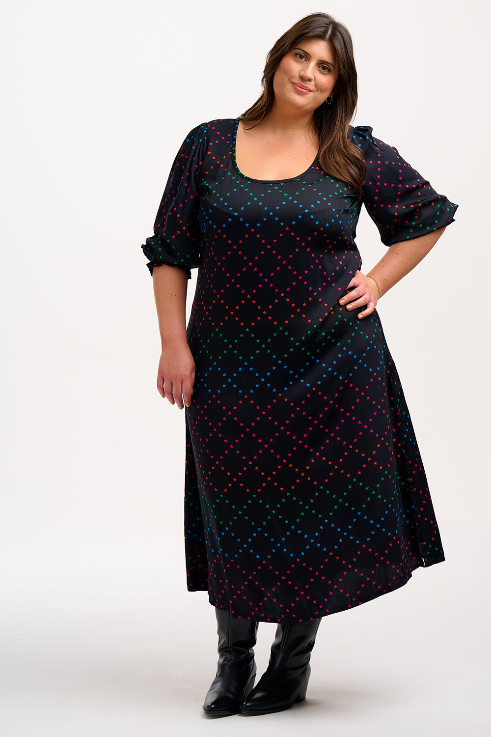 A woman standing and posing in a midi black dress with a colorful geometric star pattern on a black background, featuring a scoop neck, three-quarter sleeves, and a tie waist.