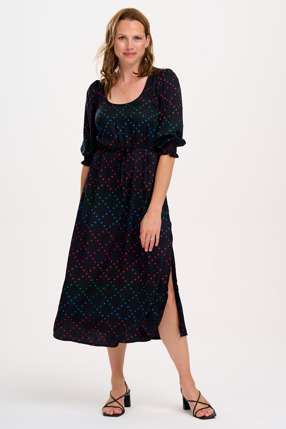 A woman standing and posing in a black and rainbow star lattice print midi dress with three-quarter length sleeves and a scoop neck.