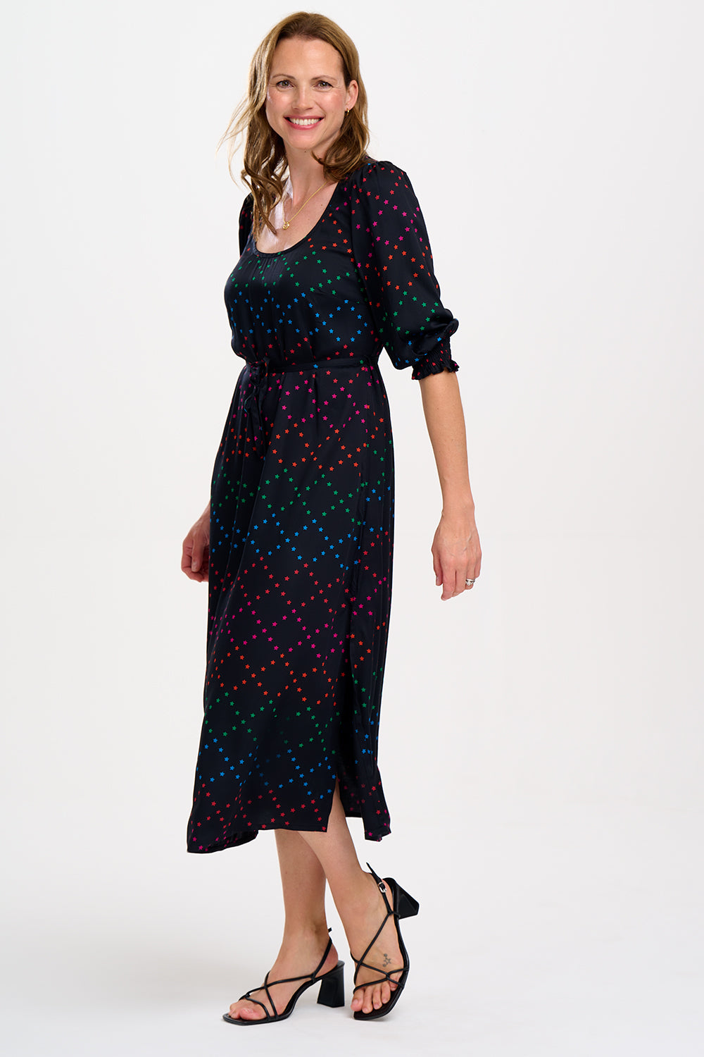 A woman standing and posing in a black and rainbow star lattice print midi dress with three-quarter length sleeves and a scoop neck.