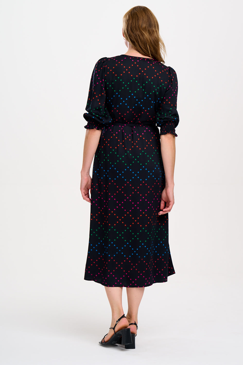 A woman standing and posing in a black and rainbow star lattice print midi dress with three-quarter length sleeves and a scoop neck.